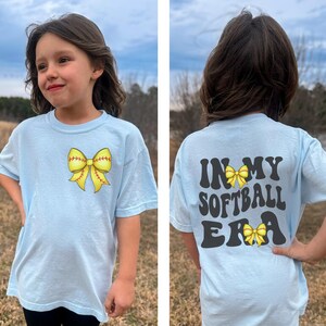 K&ouml;nnte beinhalten: Hellblaues T-Shirt mit einer gelben Softball-Schleife auf der Vorderseite und dem Text "In My Softball Era" mit einer gelben Softball-Schleife auf der R&uuml;ckseite.