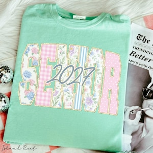 May include: A mint green t-shirt featuring the word SENIOR in large block letters with a gold outline. The letters contain floral, gingham, and striped patterns in pink, blue, and purple. The number 2027 is printed in a blue script font over the text.