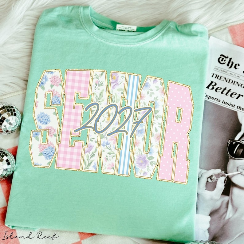 May include: A mint green t-shirt featuring the word SENIOR in large block letters with a gold outline. The letters contain floral, gingham, and striped patterns in pink, blue, and purple. The number 2027 is printed in a blue script font over the text.