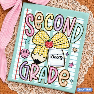 May include: A light blue notebook cover featuring the text SECOND GRADE in colorful bubble letters. The design includes a large yellow gingham bow, a pencil, a red apple, a pink smiley face, and stars. The name Kinley is printed on a white label.