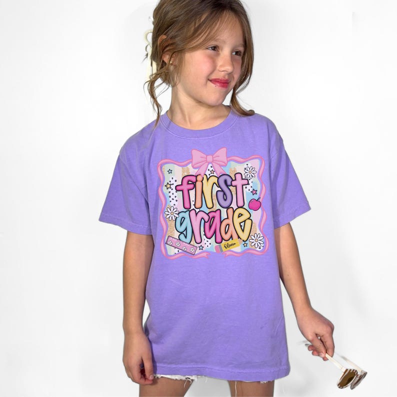 Op de afbeelding: Een lavendelkleurig T-shirt met de tekst "first grade" in kleurrijke letters. De afbeelding bevat een roze strik, sterren, bloemen en een hart. Het T-shirt wordt door een jong meisje gedragen.