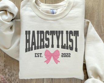 May include: A cream-colored sweatshirt with the text "HAIRSTYLIST EST. 2022" in black and a pink bow.