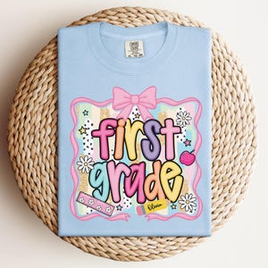 Op de afbeelding: Lichtblauw T-shirt met een kleurrijk "first grade" ontwerp. Het ontwerp bevat een roze strik, madeliefjes, sterren, een liniaal, een appel en een potlood met de naam "Olivia". Het shirt ligt op een geweven oppervlak.