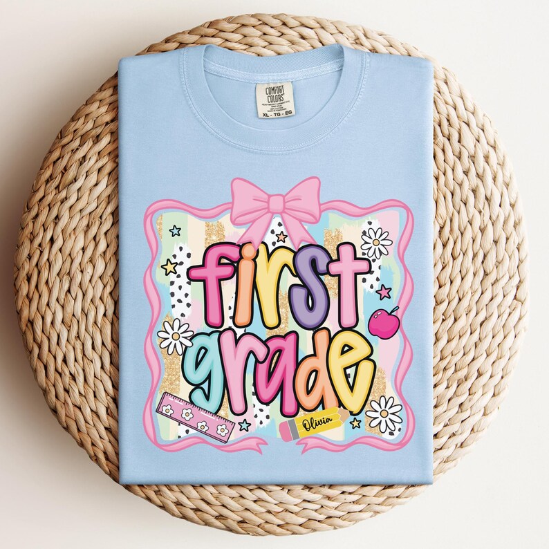 Op de afbeelding: Lichtblauw T-shirt met een kleurrijk "first grade" ontwerp. Het ontwerp bevat een roze strik, madeliefjes, sterren, een liniaal, een appel en een potlood met de naam "Olivia". Het shirt ligt op een geweven oppervlak.