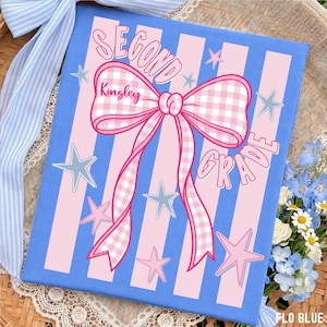 Può includere: Una stampa su tela rettangolare con uno sfondo a righe verticali azzurre e rosa. Un grande fiocco a quadretti vichy rosa e bianco è centrato con il nome Kingley scritto sopra. Il testo recita Second Grade, circondato da stelle rosa e blu.