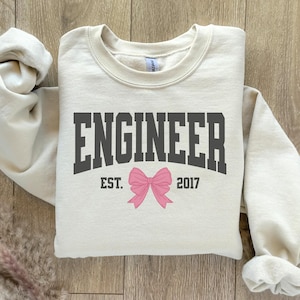 Könnte beinhalten: Ein cremefarbener Pullover mit dem Text "Engineer Est. 2017" in schwarzen Buchstaben. Eine rosafarbene Schleife befindet sich mittig unter dem Text.
