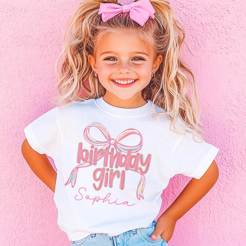 以下が含まれることがあります： 「birthday girl」の文字と「Sophia」という名前がピンク色の筆記体で書かれた白いTシャツ。カラフルなリボンデザインが特徴です。女の子は髪にピンクのリボンをつけ、笑顔です。
