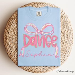 May include: A light blue t-shirt with a pink bow and the word "Dance" in pink bubble letters. Below the word "Dance" is the name "Sophia" in a cursive font. The shirt is displayed on a woven, circular surface.