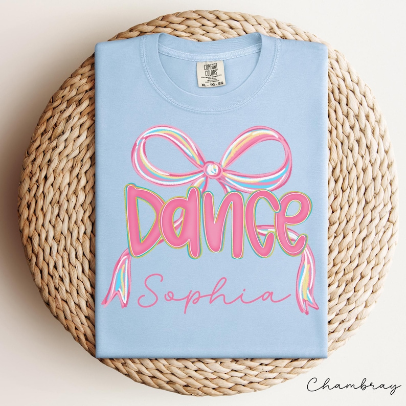 May include: A light blue t-shirt with a pink bow and the word "Dance" in pink bubble letters. Below the word "Dance" is the name "Sophia" in a cursive font. The shirt is displayed on a woven, circular surface.