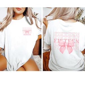 May include: White t-shirt with the text "FIFTEEN" repeated in a retro font and a pink bow on the back. The front of the shirt has the text "FIFTEEN" repeated in a retro font with the name "Mackenzie" underneath.