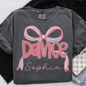 May include: Dark grey sweatshirt featuring a pink bow and the word "DANCE" in pink letters with a yellow outline. The name "Sophia" is written below the word "DANCE" in a cursive font. The bow has rainbow-coloured ribbons.