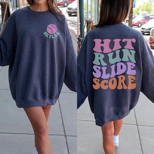 May include: A dark gray sweatshirt with a baseball graphic on the front and the text "Skylar". The back of the sweatshirt has the words "Hit Run Slide Score" in a retro font. The sweatshirt is a comfortable, oversized fit.
