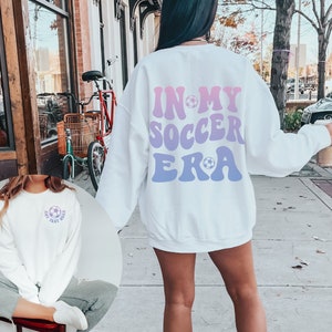 May include: White sweatshirt with the text "In My Soccer Era" in a retro font with a football graphic. The text is in pink and blue.