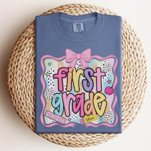 Op de afbeelding: Een blauw Comfort Colors T-shirt met een kleurrijke "first grade" afbeelding. De afbeelding bevat een roze strik, bloemen, sterren en een potlood met de naam "Olivia". Het shirt wordt gepresenteerd op een geweven, ronde placemat.