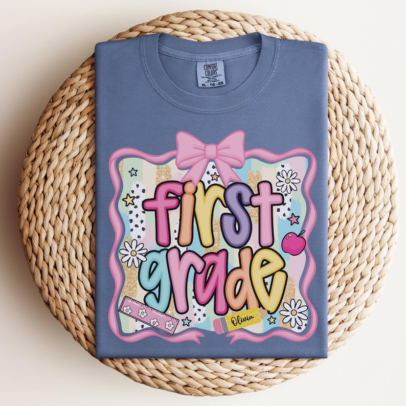 Op de afbeelding: Een blauw Comfort Colors T-shirt met een kleurrijke "first grade" afbeelding. De afbeelding bevat een roze strik, bloemen, sterren en een potlood met de naam "Olivia". Het shirt wordt gepresenteerd op een geweven, ronde placemat.