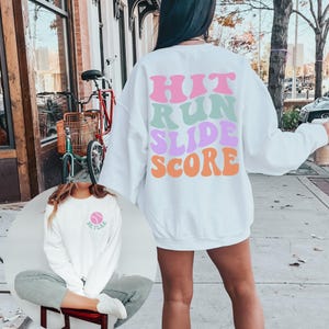 May include: White sweatshirt with the words "HIT RUN SLIDE SCORE" in pink, green, purple, and orange. A smaller image shows a white sweatshirt with a baseball graphic and the name "Skylar".