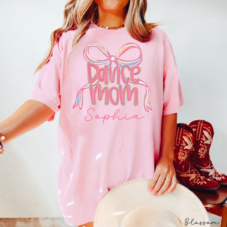 May include: A light pink t-shirt with the words "Dance Mom" and the name "Sophia" in a script font. The design includes a colorful bow and ribbons. The shirt is paired with a white hat and red cowboy boots.