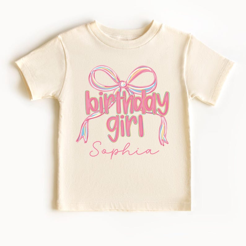 以下が含まれることがあります： カラフルなリボンとピンク色の「birthday girl」の文字、下に「Sophia」の名前が入ったクリーム色のTシャツ。デザインは半袖シャツの前面中央に配置されています。