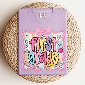 Op de afbeelding: Een lila t-shirt met een kleurrijke "first grade" afbeelding. Het ontwerp bevat een roze strik, madeliefjes, sterren, een appel en een liniaal. De naam "Olivia" staat op een potlood.