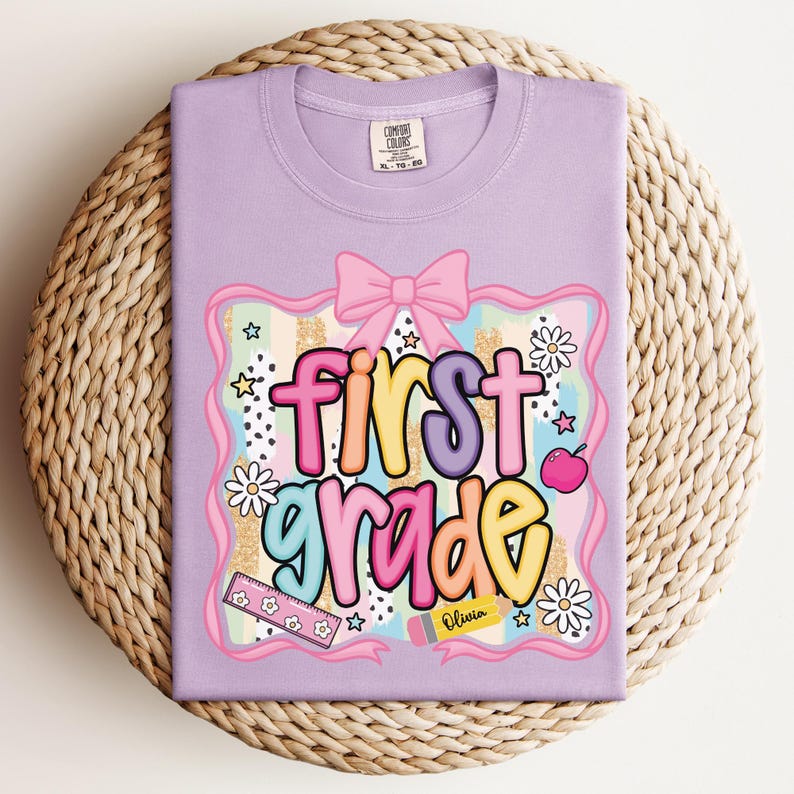 Op de afbeelding: Een lila t-shirt met een kleurrijke "first grade" afbeelding. Het ontwerp bevat een roze strik, madeliefjes, sterren, een appel en een liniaal. De naam "Olivia" staat op een potlood.