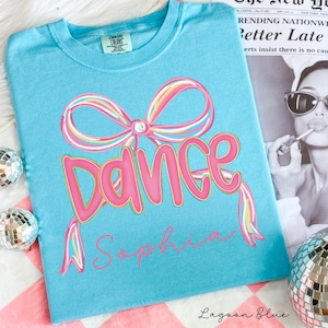 May include: Lagoon blue t-shirt with a pink "Dance" graphic and a bow design. The word "Sophia" is written below the word "Dance". The shirt is made of a soft material and has a crew neck.