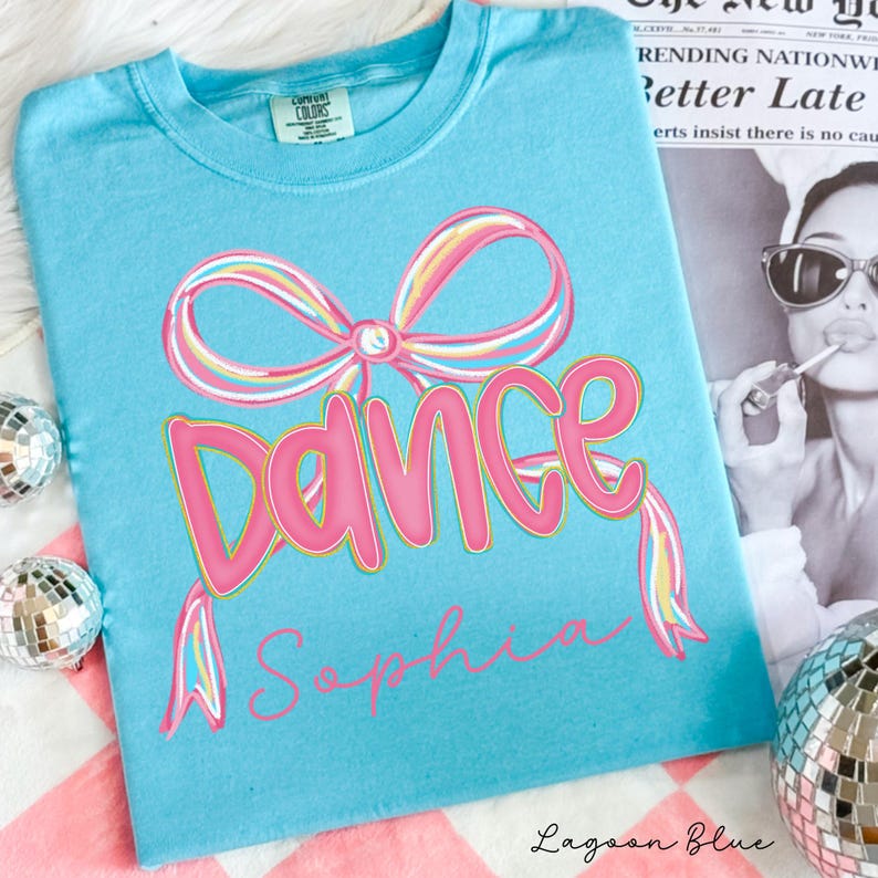 May include: Lagoon blue t-shirt with a pink "Dance" graphic and a bow design. The word "Sophia" is written below the word "Dance". The shirt is made of a soft material and has a crew neck.