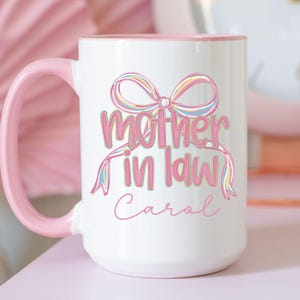 May include: White ceramic mug with a pink handle and rim. The mug features the text "mother in law" in pink script with a rainbow bow design. The name "Carol" is written below the text.