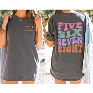 Pode incluir: Uma camiseta cinza escura com um gráfico de dançarinos na frente e o texto "FIVE SIX SEVEN EIGHT" em uma fonte retrô colorida nas costas. A camiseta é feita de um material macio e confortável.