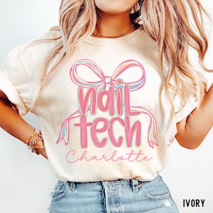 Op de afbeelding: Ivoor kleurig T-shirt met de tekst "Nail Tech Charlotte" in roze letters, geaccentueerd door een pastelkleurig strik ontwerp. Het shirt is een casual stijl, geschikt voor dagelijks gebruik.