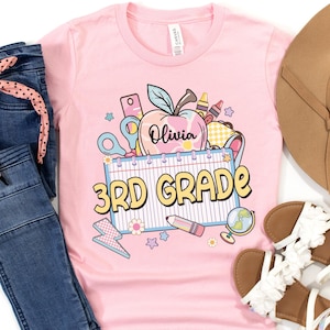 以下が含まれることがあります： 明るいピンク色のTシャツ。学校をテーマにしたカラフルなグラフィックが特徴で、リンゴ、ハサミ、クレヨン、そして「3rd Grade」と「Olivia」の文字が描かれています。青いジーンズと白いサンダルとコーディネートされています。