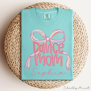 May include: A light blue t-shirt with a pink bow and the words "Dance Mom" in pink script. The name "Sophia" is written below. The shirt is folded on a woven, circular surface.