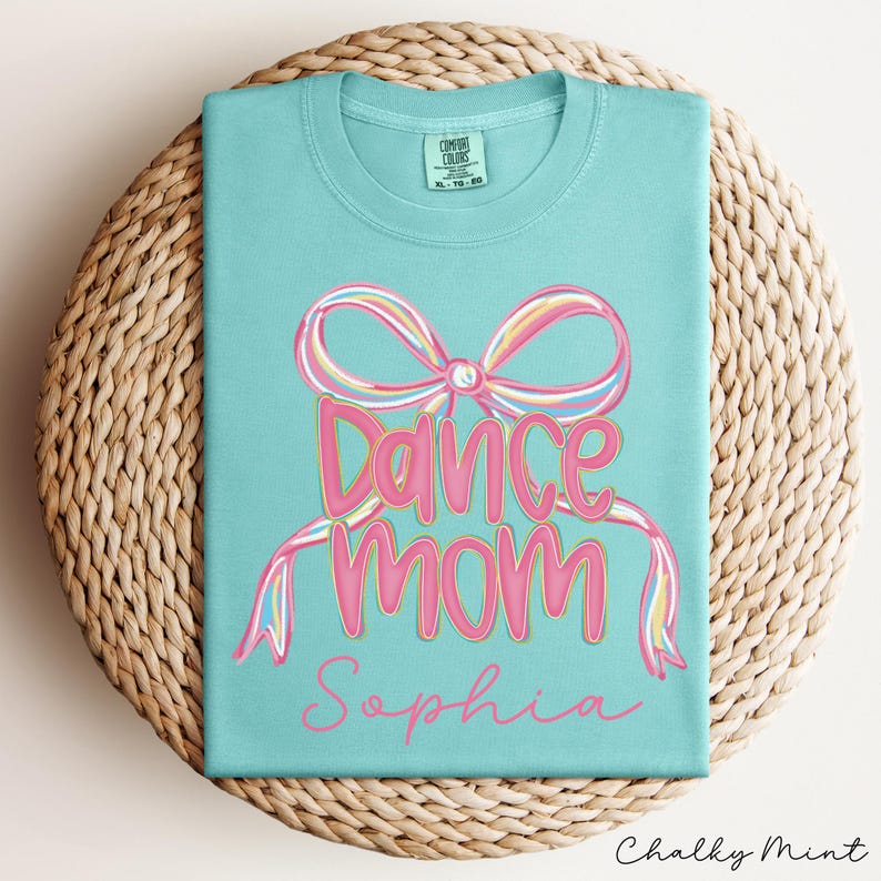 May include: A light blue t-shirt with a pink bow and the words "Dance Mom" in pink script. The name "Sophia" is written below. The shirt is folded on a woven, circular surface.