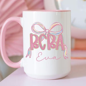 Könnte beinhalten: Weißer Keramikbecher mit rosa Griff und Rand. Der Becher zeigt den Text "BCBA" in Rosa, mit einem bunten Schleifendesign über den Buchstaben. Der Name "Eva" ist darunter in Schreibschrift geschrieben. Der Becher steht auf einer rosa Oberfläche.