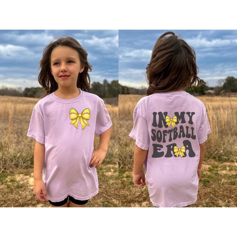 K&ouml;nnte beinhalten: Ein lavendelfarbenes T-Shirt mit einer gelben Softball-Schleife auf der Vorderseite und dem Text "In My Softball Era" mit einer gelben Softball-Schleife auf der R&uuml;ckseite.