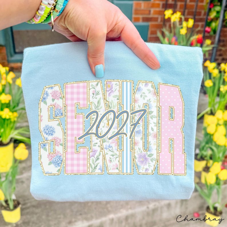 May include: A light blue t-shirt featuring the word SENIOR in large block letters with varied patterns including floral prints, pink gingham, and blue stripes. The number 2027 is printed in a cursive grey font across the center of the letters.