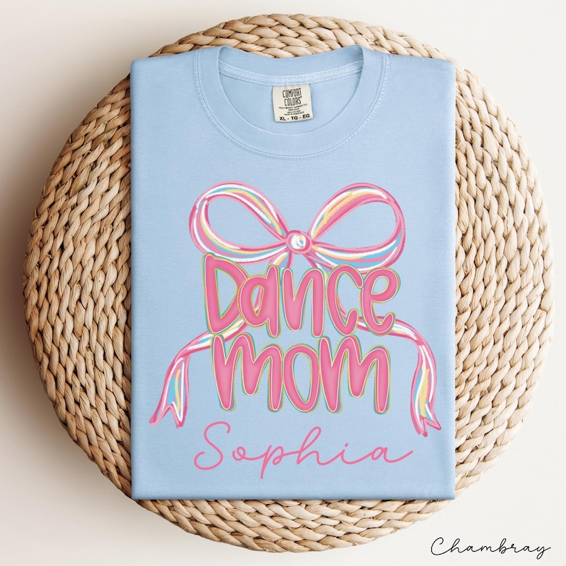 May include: A light blue t-shirt with a colorful graphic that says "Dance Mom Sophia". The design features a large bow and the words "Dance Mom" in pink with a green outline. The name "Sophia" is written in a cursive font.