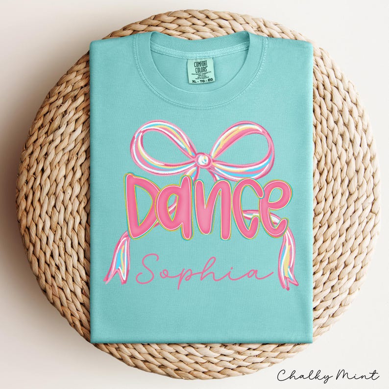 May include: A turquoise t-shirt with a pink and rainbow bow design and the word "Dance" in pink, with the name "Sophia" in cursive. The shirt is displayed on a woven, circular placemat.