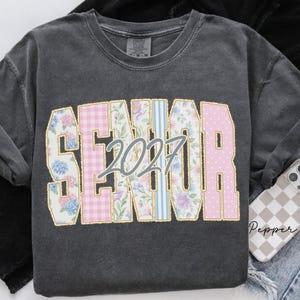 May include: A charcoal gray crewneck t-shirt featuring the word SENIOR in large block letters with varied patterns including floral, pink gingham, and blue stripes. The number 2027 is printed in a light blue script font across the center of the text.