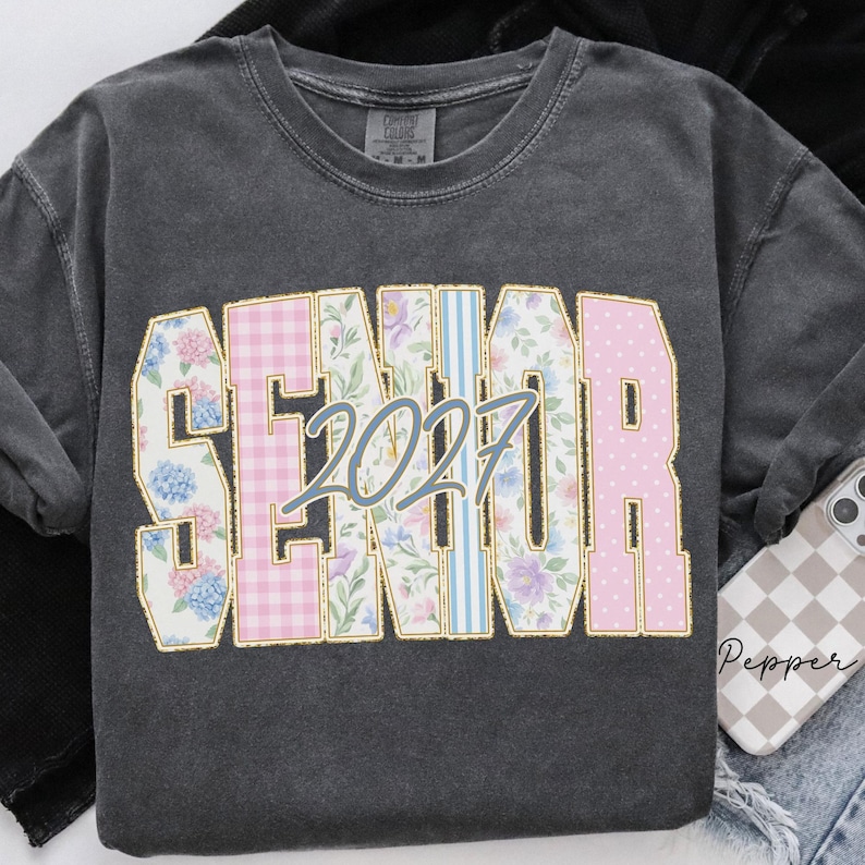 May include: A charcoal gray crewneck t-shirt featuring the word SENIOR in large block letters with varied patterns including floral, pink gingham, and blue stripes. The number 2027 is printed in a light blue script font across the center of the text.
