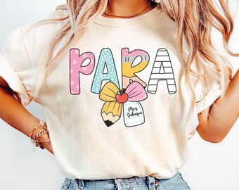 Könnte beinhalten: Cremefarbenes T-Shirt mit dem Wort "PARA" in bunten, verspielten Buchstaben. Das Design umfasst einen Bleistift, eine Schleife und ein Etikett mit der Aufschrift "Mrs. Johnson". Das Shirt ist ein lässiger Stil.