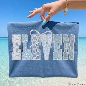 May include: A blue t-shirt with the word "ELEVEN" in white, light blue, and pearl embellishments. The letters have various patterns, including starfish, seashells, and wave designs. The shirt is held up against a beach backdrop.