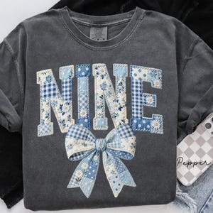 Könnte beinhalten: Ein graues Sweatshirt mit dem Wort "NINE" in einem Patchwork-Design appliziert. Die Buchstaben zeigen blaue und weiße Blumen-, Tupfen- und Vichy-Muster. Eine passende Schleife befindet sich unter dem Wort.