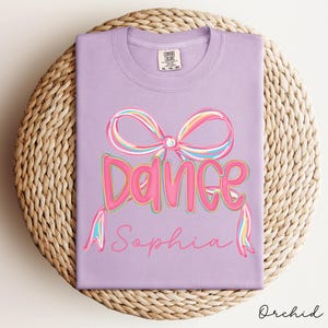 May include: Orchid-colored t-shirt with a large pink bow and the word "Dance" in pink, outlined in a darker pink. Below the word "Dance" is the name "Sophia" in a cursive font. The shirt is folded on a woven mat.