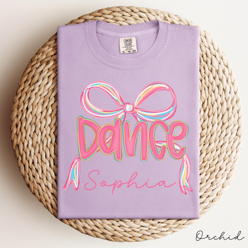 May include: Orchid-colored t-shirt with a large pink bow and the word "Dance" in pink, outlined in a darker pink. Below the word "Dance" is the name "Sophia" in a cursive font. The shirt is folded on a woven mat.