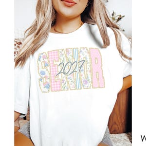 May include: A white short-sleeve t-shirt featuring the word SENIOR in large block letters with a gold outline. The letters contain floral, plaid, and striped patterns in pink, blue, and white. The number 2027 is printed in gray over the letters.