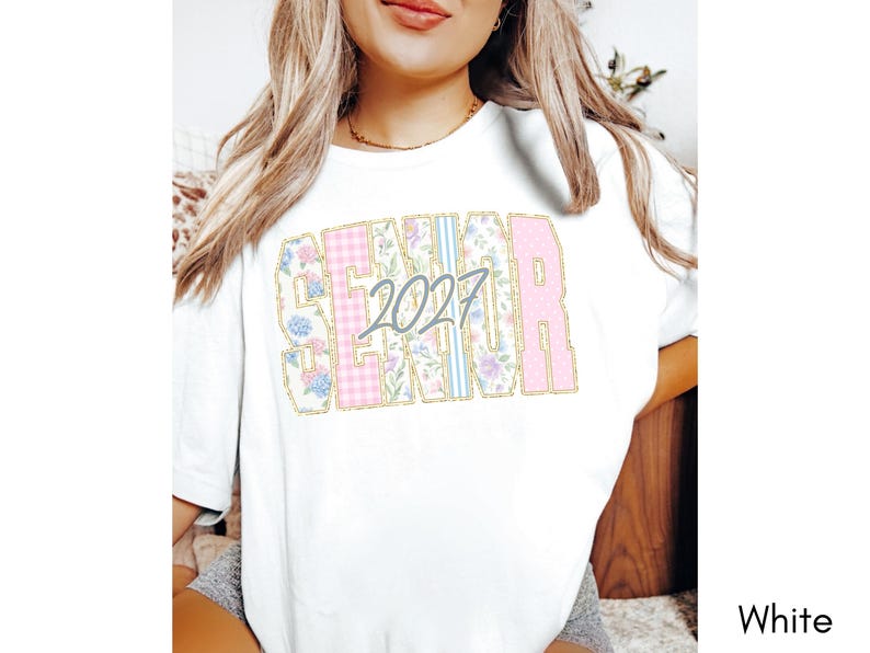 May include: A white short-sleeve t-shirt featuring the word SENIOR in large block letters with a gold outline. The letters contain floral, plaid, and striped patterns in pink, blue, and white. The number 2027 is printed in gray over the letters.