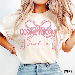 May include: Ivory-colored t-shirt featuring a pink bow design with the word "Cosmetology" in pink script. Below the word is the name "Sophia" in a cursive font. The shirt is a casual style.