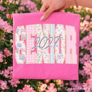 May include: A bright pink t-shirt featuring the word SENIOR in large block letters with varied patterns including floral, gingham, stripes, and polka dots. The text 2027 is layered over the center in a light blue script font with a gold outline.