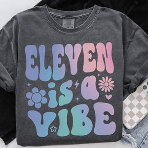 Op de afbeelding: Donkergrijze sweatshirt met de tekst "ELEVEN is a VIBE" in een kleurrijk retro-lettertype. Het ontwerp bevat bloemen, sterren en een bliksemschicht. De sweatshirt heeft een ronde hals en lange mouwen.