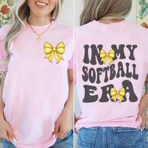 K&ouml;nnte beinhalten: Pinkes T-Shirt mit einer gelben Softball-Schleife auf der Vorderseite und dem Text "In My Softball Era" mit einer gelben Softball-Schleife auf der R&uuml;ckseite.